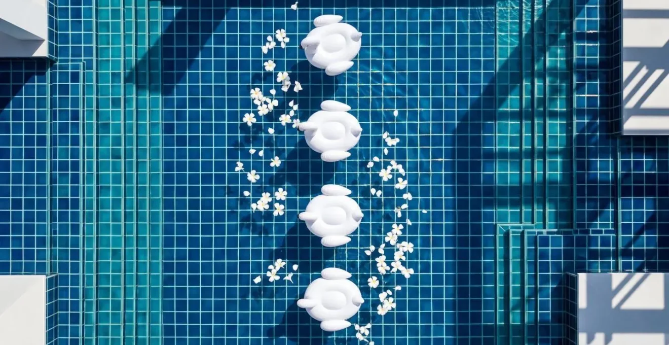Vista dall'alto di piscina blu profondo con fenicotteri gonfiabili bianchi disposti simmetricamente e petali bianchi galleggianti, ombre architettoniche geometriche